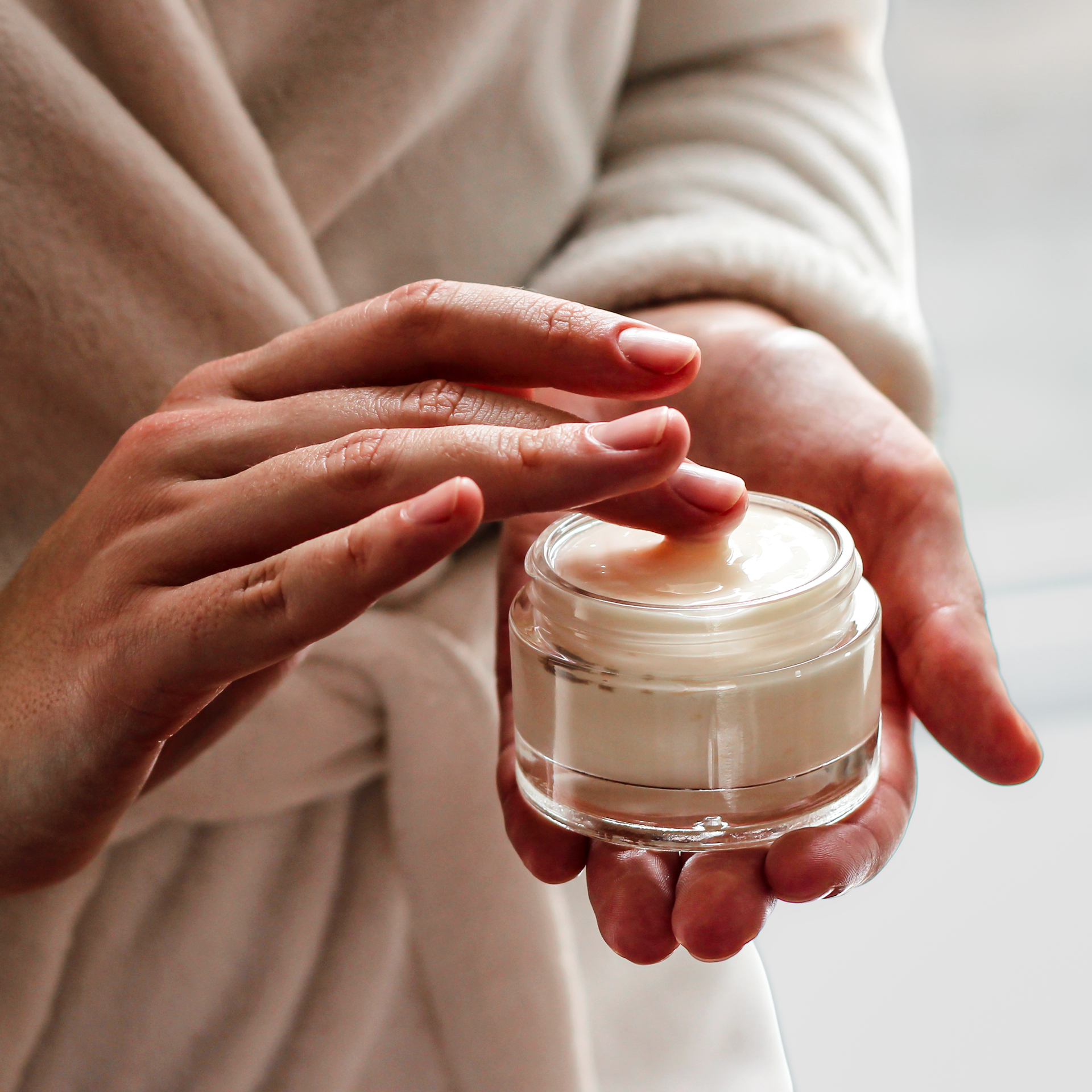 Two hands holding a small glass jar of white cream, one fingertip lifting a little from the surface.