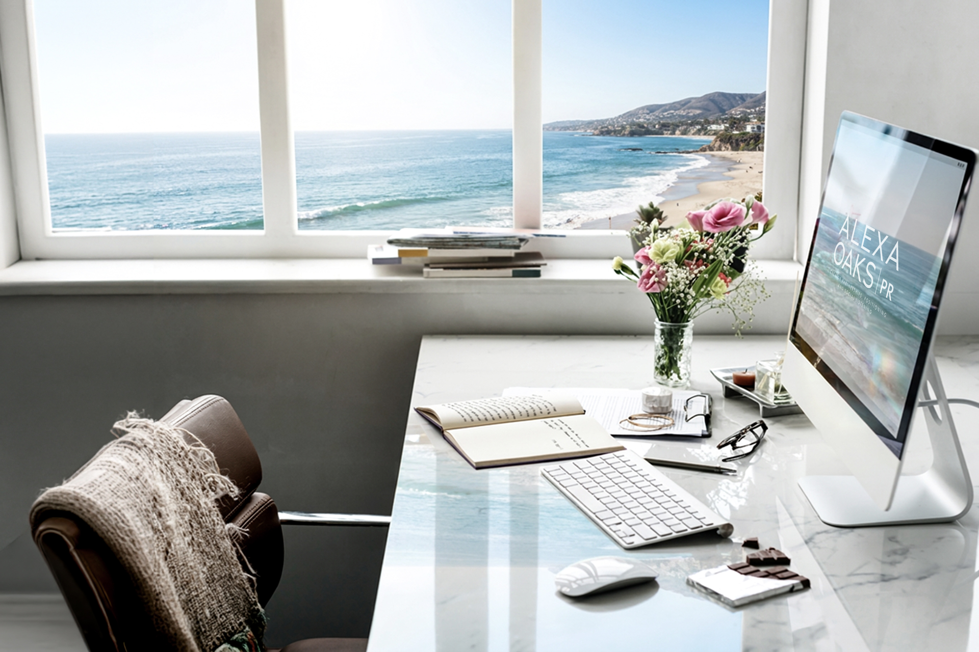 An empty desk in a bright office above the beach at Laguna—open notebook, keyboard, reading glasses, fresh flowers, and a monitor showing the Alexa Oaks PR logo. Nobody is in the chair.