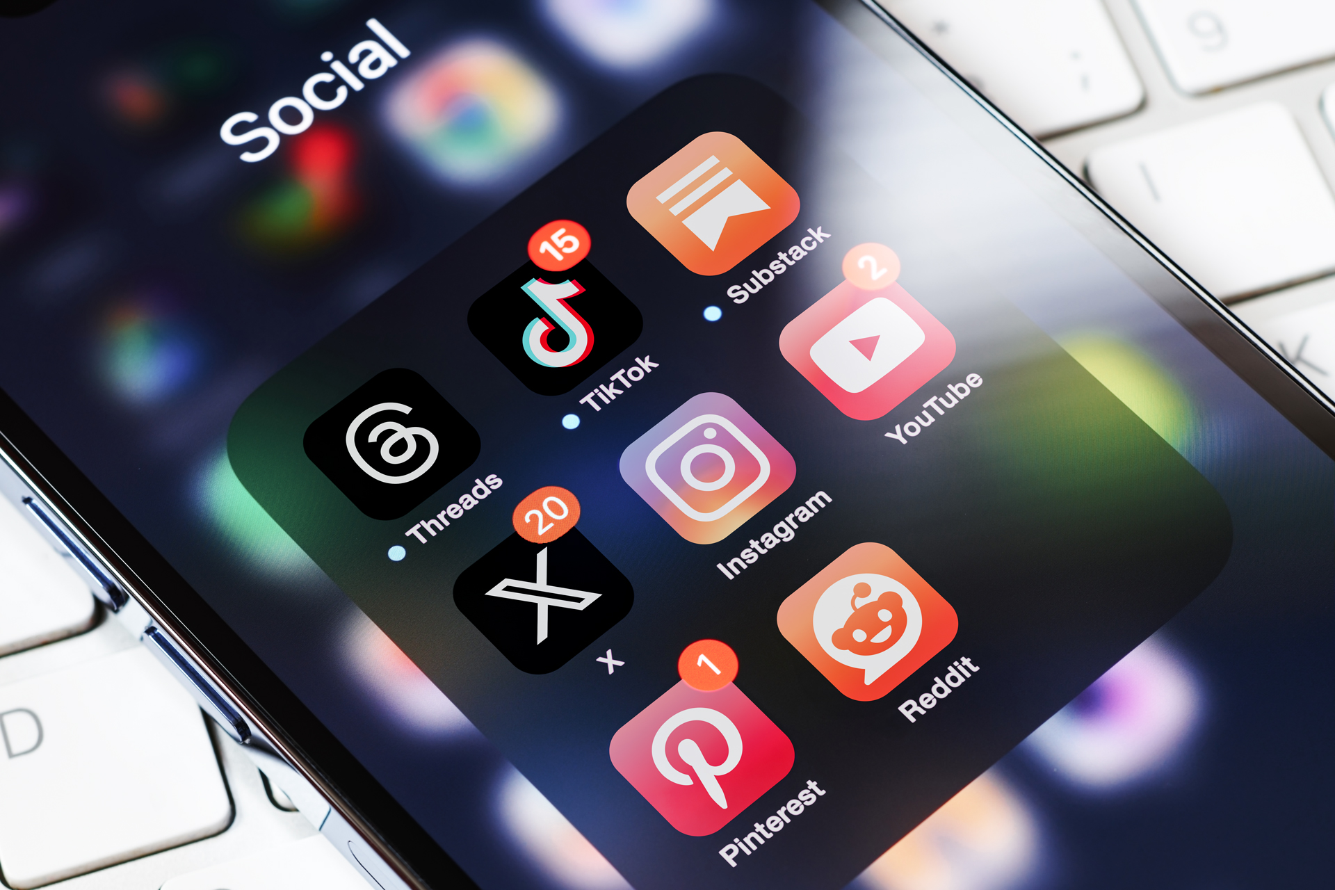 A phone resting on a keyboard, open to a folder of social apps—Threads, TikTok, Substack, Instagram, YouTube, X, Pinterest and Reddit—several carrying unread notification counts.