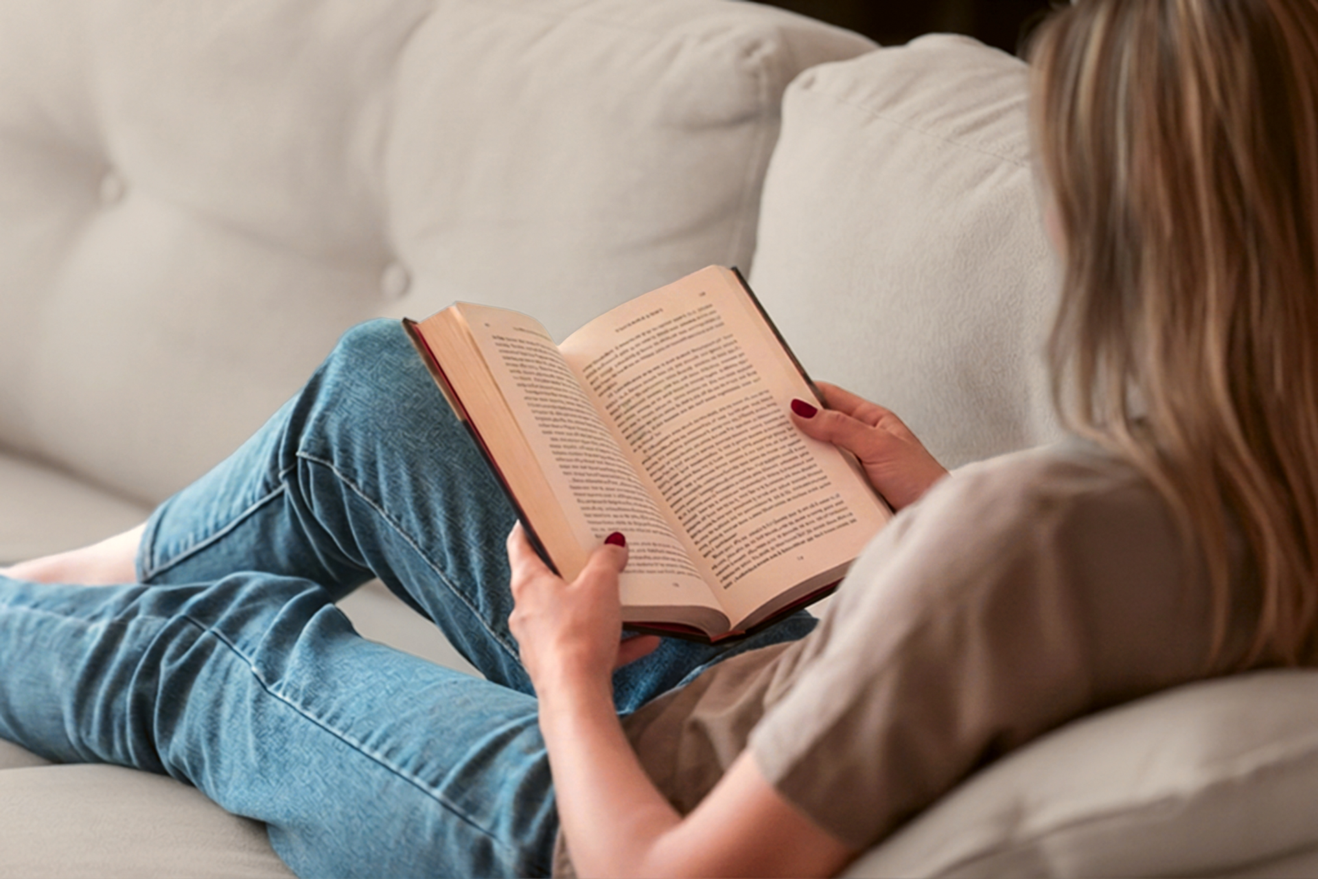 Alexa reading a hardback book on a sofa with her feet up, in afternoon light.